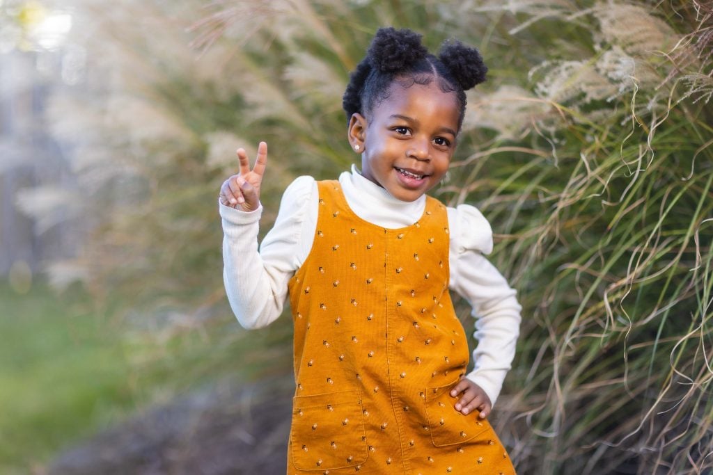 A young child stands outdoors next to tall plants, wearing a yellow dress and white shirt, holding up a peace sign with one hand while the other is on their hip, smiling at the camera in this charming family portrait.