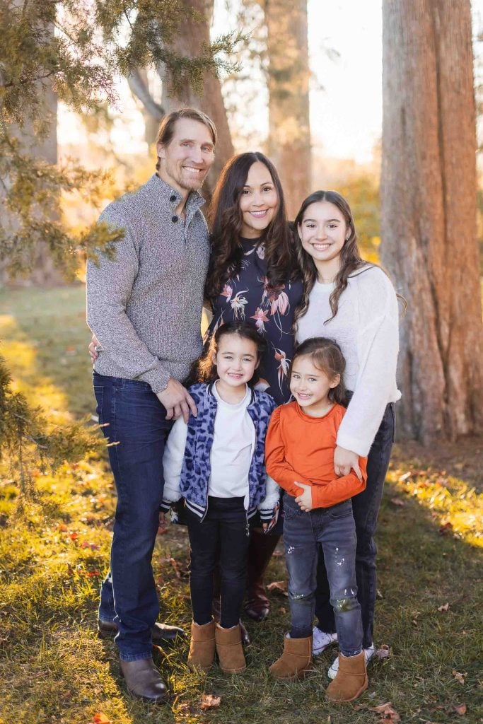 A family of five posing outdoors in a sunlit forested area, with two adults standing behind three young children who are smiling and dressed in casual clothing; it feels like a natural, candid snapshot reminiscent of timeless portraits taken at Belair Mansion.