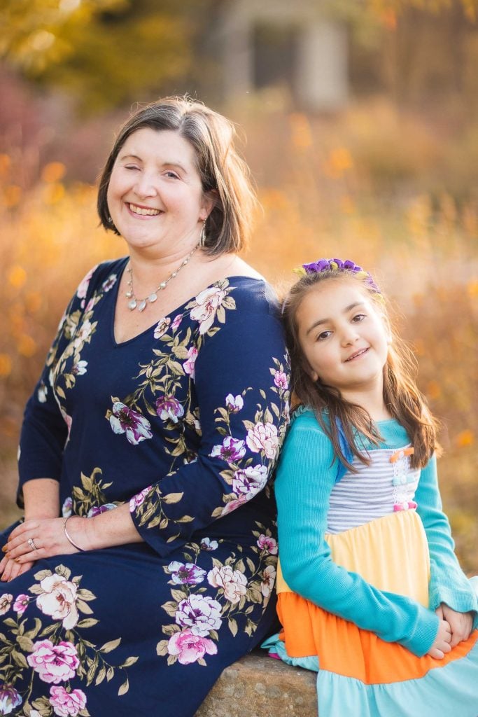 A woman in a floral dress sits and smiles beside a young girl in a colorful dress with a purple headband, both enjoying an outdoor autumn setting at Greenspring Garden in Alexandria, Virginia.