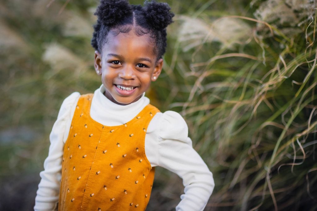 A young girl with puffballstyle buns smiles at the camera in this charming family portrait, wearing a yellow dress over a white longsleeve shirt, standing outdoors with greenery in the background.