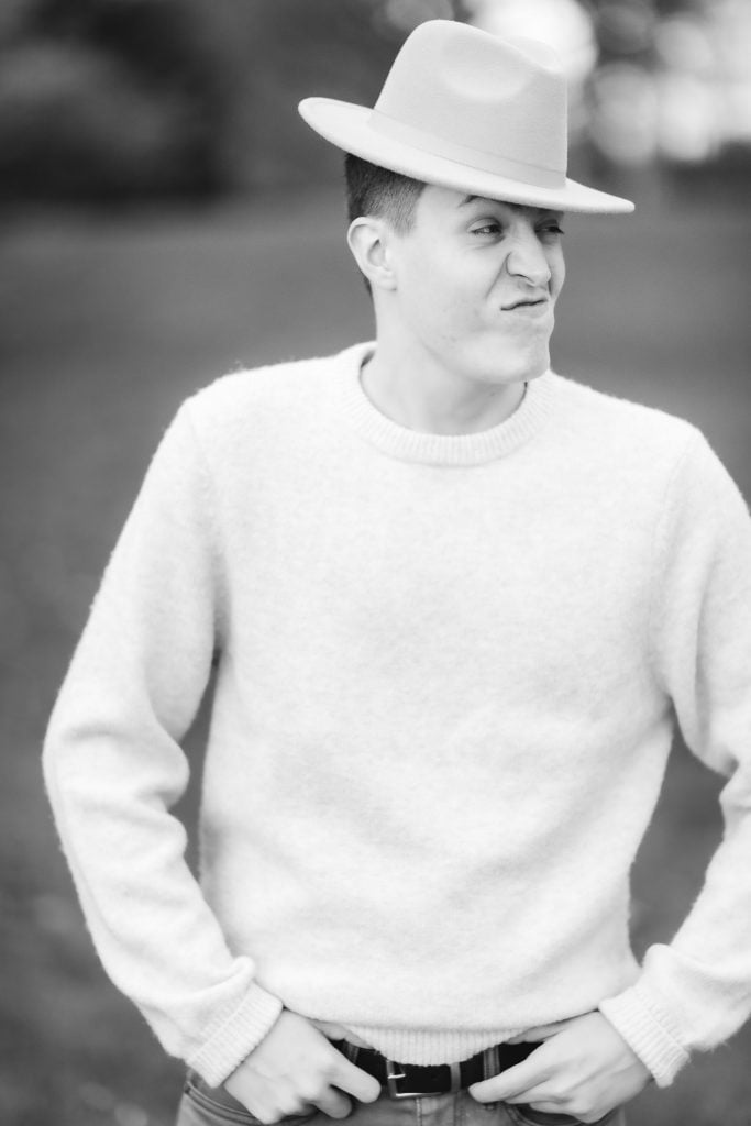 Black and white portrait of a person outdoors, wearing a sweater and a hat, making a playful facial expression.