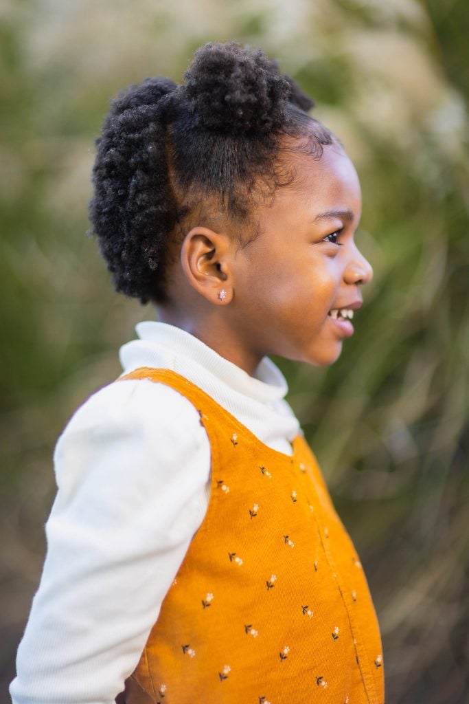 Young child in profile wearing a white turtleneck under a yellow dress, standing outdoors with a blurry natural background. The portrait captures a tender family moment bathed in soft, natural light.