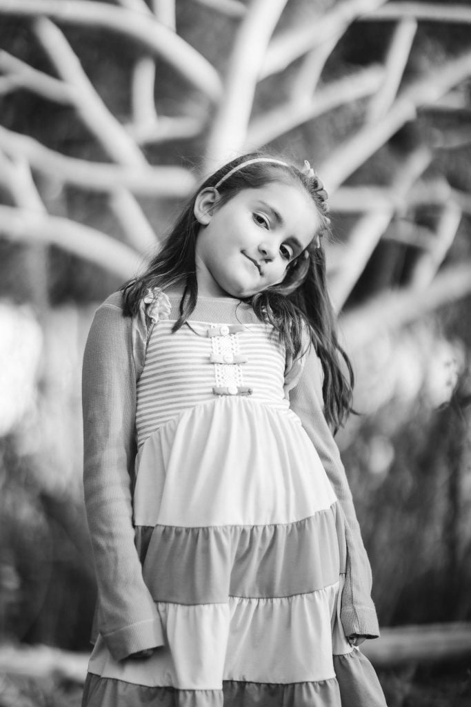 A young girl stands outdoors in front of a tree at Greenspring Garden. She has long hair and is wearing a striped dress with long sleeves. The photo, captured in black and white, evokes a timeless charm.