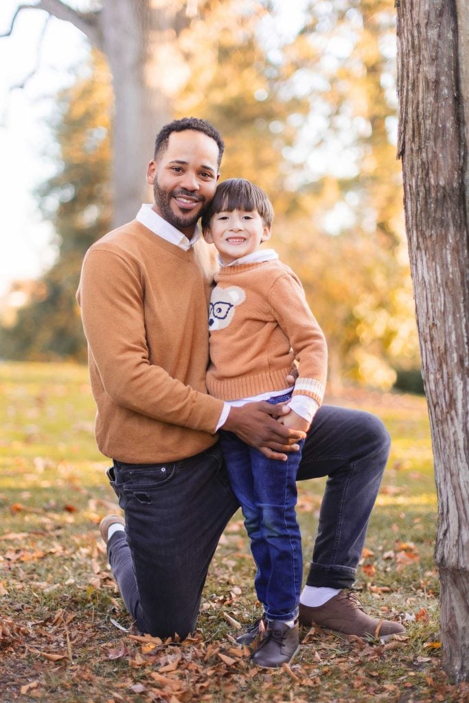 A man kneels on one knee next to a tree, holding a young boy with one arm. Both are wearing brown sweaters and smiling at the camera. The background shows a grassy area with autumn leaves, perfect for capturing timeless portraits that could adorn the walls of any home, including Belair Mansion.