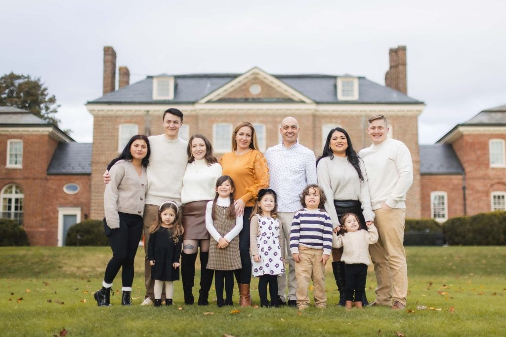 A group of eleven people, including adults and children, stand together on a grassy lawn in front of the stately Belair Mansion, capturing a charming family portrait.