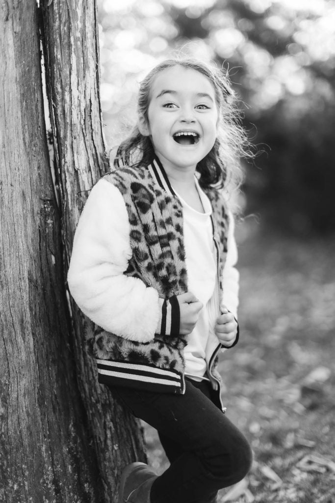 A smiling young girl stands next to a tree, wearing a furry jacket with a leopard print pattern. She appears to be laughing and is positioned in an outdoor setting near Belair Mansion. The image is in black and white.