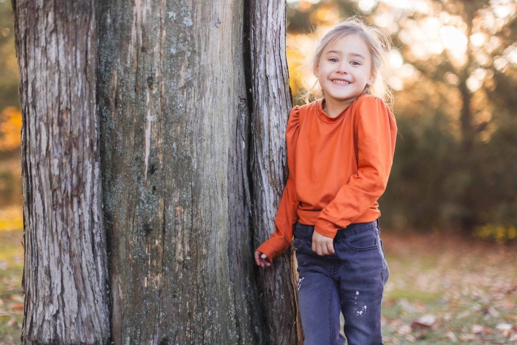 A young girl with a smile stands next to a large tree wearing an orange longsleeve shirt and dark pants. The background features an outdoor setting with sunlight filtering through the trees, perfect for capturing timeless portraits reminiscent of family gatherings at Belair Mansion.