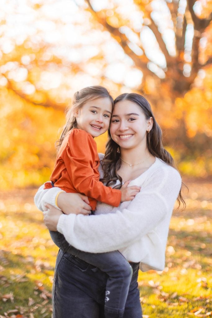 A woman in a white sweater holds a smiling child in an orange shirt. They are standing outdoors with autumn foliage in the background, reminiscent of timeless portraits often seen at Belair Mansion.