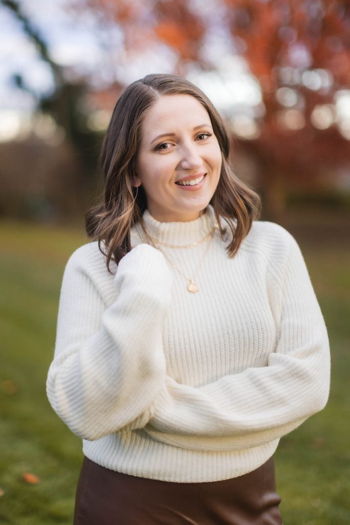 A portrait of a woman with shoulderlength brown hair wearing a white sweater and brown skirt smiles outdoors with autumn foliage in the background, creating a timeless memory at Belair Mansion.