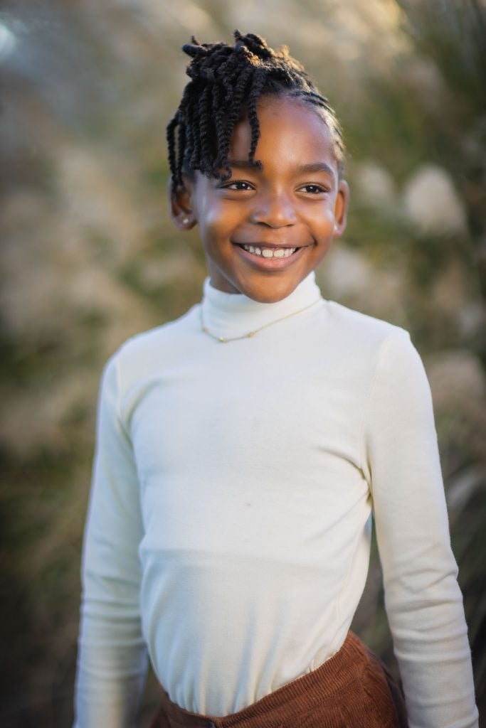 A young girl with braided hair stands outdoors, smiling and wearing a lightcolored turtleneck shirt and brown pants. This charming portrait captures her joy amid the blurred greenery in the background.