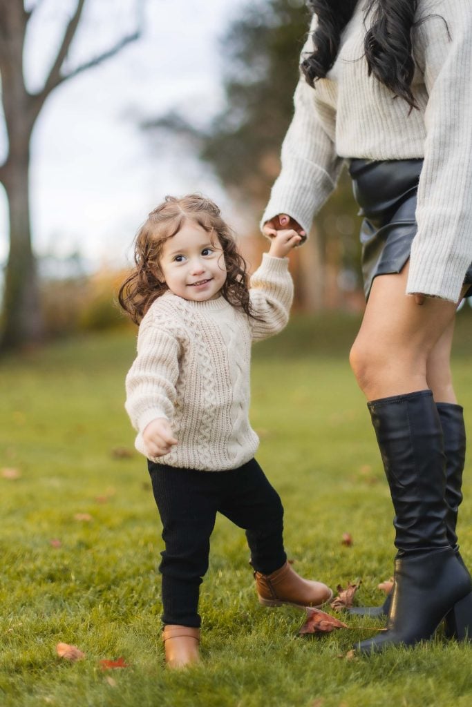 A young child with curly hair, wearing a beige sweater, holds an adult's hand and smiles while standing on the grass outdoors. The portrait captures the family moment perfectly as the adult, dressed in a cream sweater and black skirt with kneehigh boots, enjoys a day out at Belair Mansion.