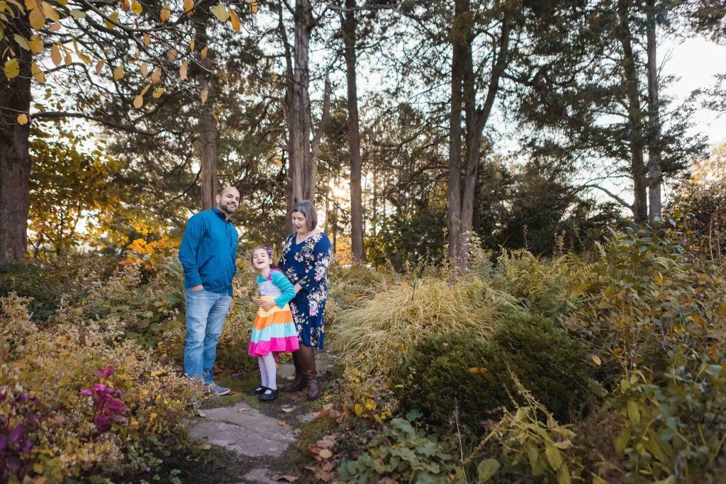 A man, woman, and young girl stand together on a stone path in Greenspring Garden with trees and plants in the background. The girl wears a colorful dress, while the adults wear blue tops. This charming portrait captures their moment in the serene beauty of Virginia.