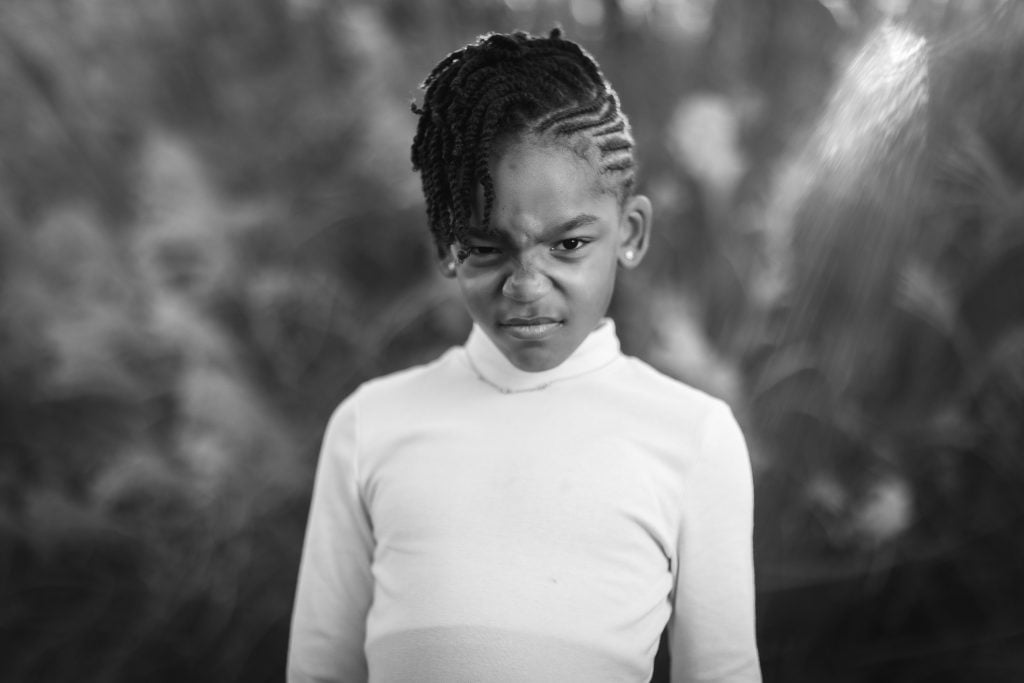 Black and white family portrait of a young girl with braided hair and a sideswept hairstyle, wearing a longsleeve top, making a scrunchedup facial expression, and standing against a blurred natural background.