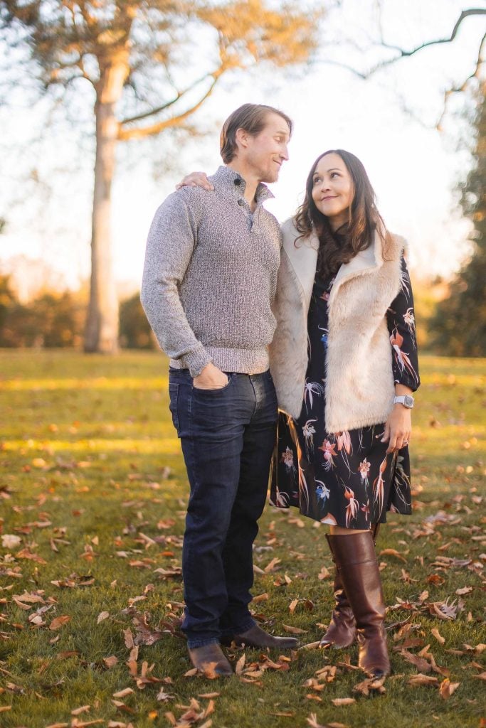 A man and woman stand on a grassy, leafstrewn area with trees in the background, smiling and looking at each other. The man wears a grey sweater and jeans, while the woman is in a dress, vest, and boots. This could be an ideal spot for extended family portraits at Belair Mansion.