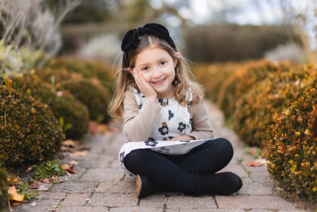 A smiling girl with a hand on her cheek sits crosslegged on a pathway bordered by neatly trimmed bushes at Belair Mansion. She is wearing a floral dress, black tights, and a black headband. This portrait captures the serene beauty of family moments in an elegant setting.