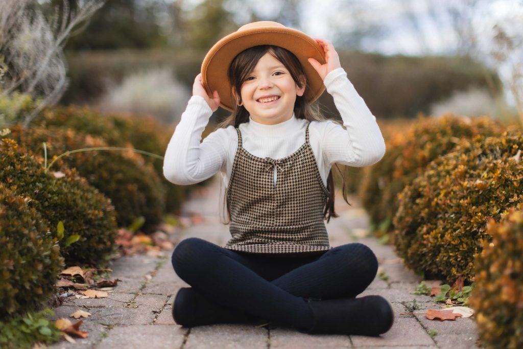 A young girl sits crosslegged on a brick pathway in front of Belair Mansion, smiling and adjusting a tan hat with both hands. She is wearing a white longsleeve shirt, patterned dress, and black leggings. This charming portrait captures her joy as the surrounding bushes blur into the background.