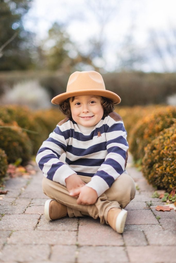 A child with curly hair sits crosslegged on a paved path, wearing a striped sweater, tan pants, tan shoes, and a tan hat. The background features blurred foliage and hedges, making it look like a serene portrait taken at Belair Mansion.