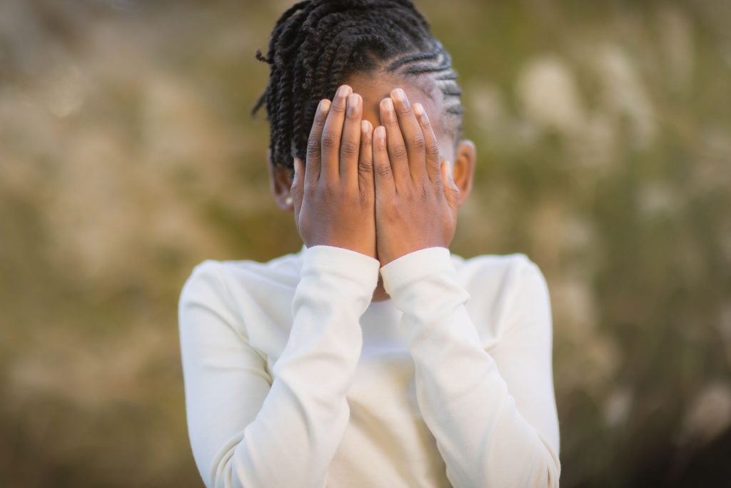 A child stands outdoors covering their face with both hands, wearing a white longsleeve shirt, capturing a candid family moment.