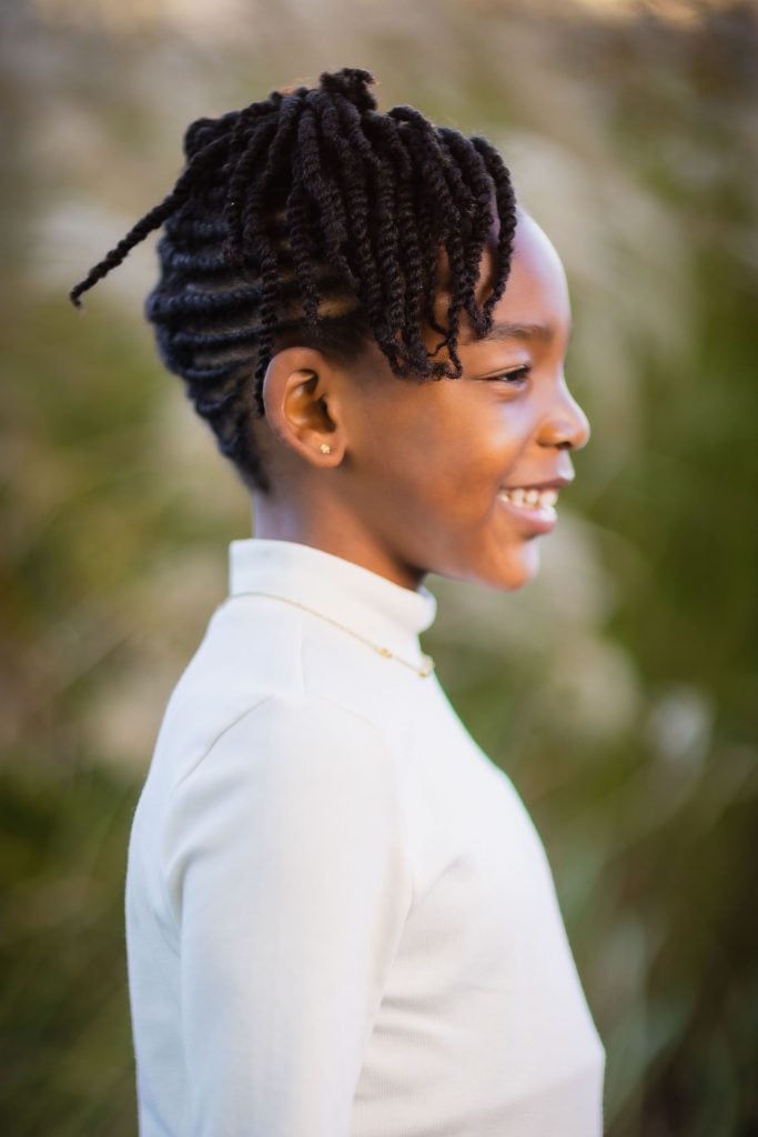 Profile of a smiling child with braided hair wearing a white shirt, standing outdoors against a blurred green background. This heartwarming portrait captures the essence of family joy and togetherness.