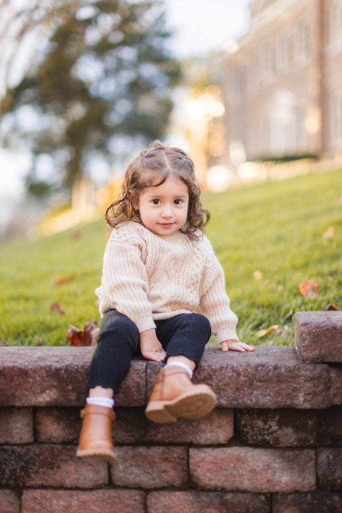 A young child with curly hair, wearing a beige sweater, black pants, and brown boots, sits on a stone ledge in an outdoor setting with green grass and trees in the background—a perfect family portrait at the Belair Mansion.