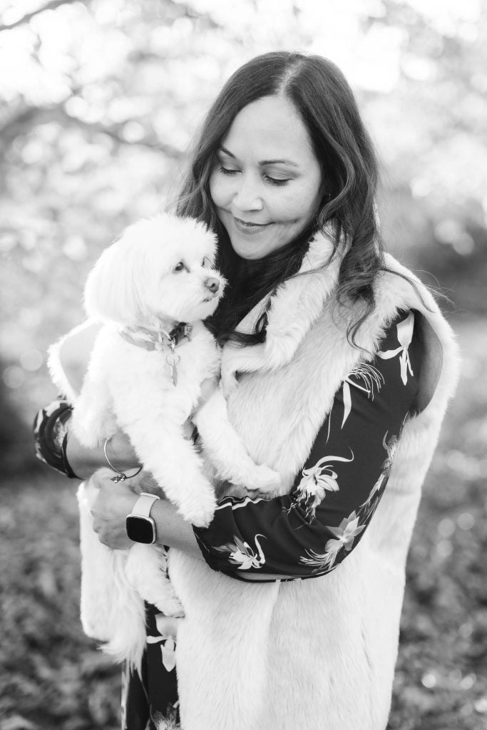 A woman wearing a furtrimmed vest holds a small white dog while smiling outdoors in a black and white photo, capturing the essence of elegant portraits often seen at the Belair Mansion.