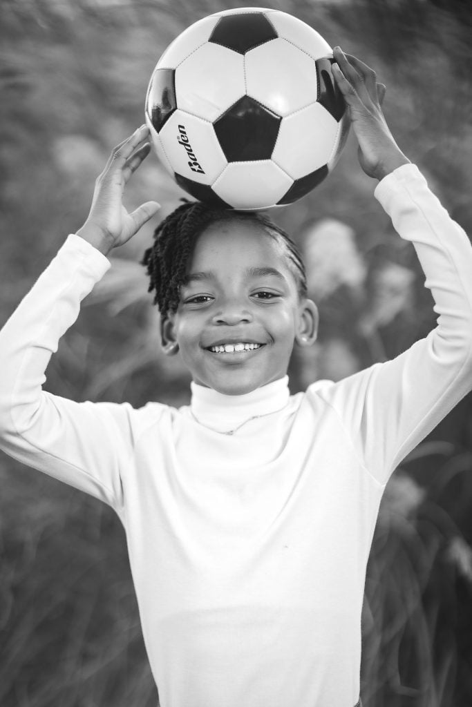 A black and white portrait of a child smiling and holding a soccer ball above their head with both hands. The child, wearing a longsleeve shirt, radiates joy, capturing the essence of family fun.