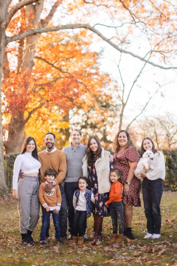 A group of seven adults and three children pose outdoors on a grassy area with autumn trees in the background at Belair Mansion. One person is holding a small white dog. Everyone is smiling and dressed in casual fall attire, capturing a perfect extended family moment.