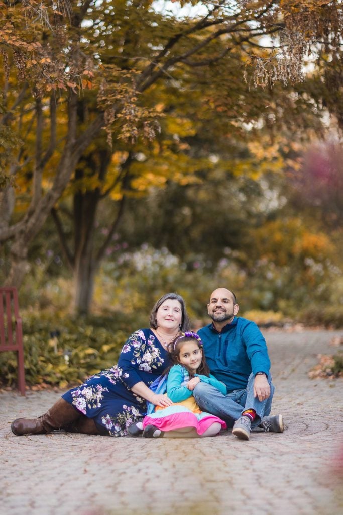 A family of three, consisting of a woman, a man, and a young girl, sits on a stone path in an Alexandria park with autumn foliage in the background—capturing the perfect Virginia portrait.