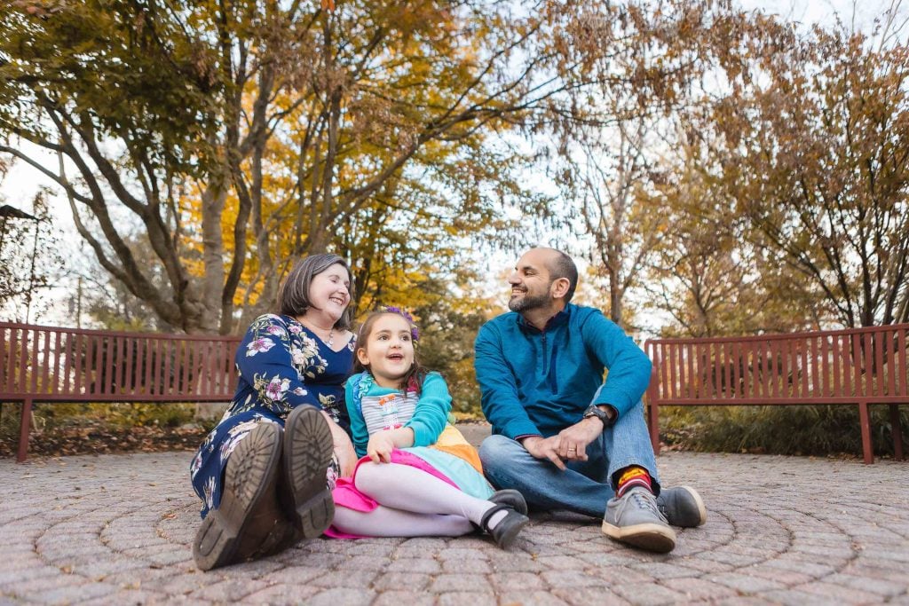 A family of three sits on a circular stone path in the enchanting Greenspring Garden. The child is in the middle, flanked by two adults. Trees with autumn leaves create a picturesque backdrop, reminiscent of Alexandria's serene charm.