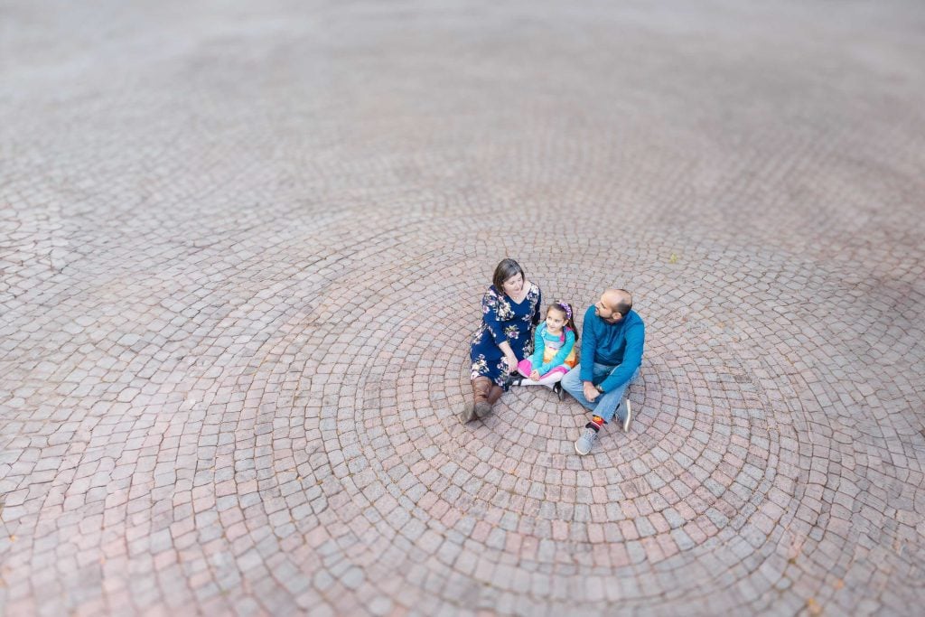 A family of three enjoys a serene moment together on the circular, patterned brick pavement at Greenspring Garden in Virginia, viewed from above.