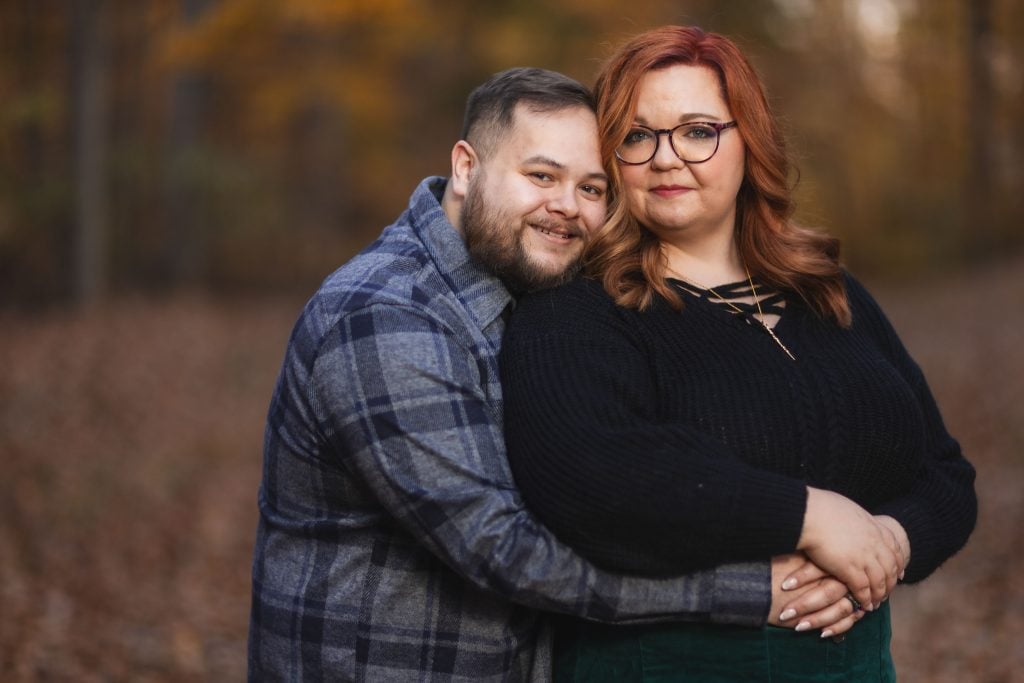 A couple stands in an autumnal forest at dusk, with the man in a plaid shirt hugging the woman in a black sweater from behind. Both are looking at the camera. The moody atmosphere adds a touch of magic to their embrace.