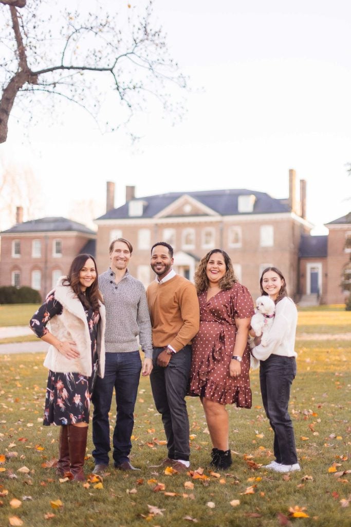 Five people and a small white dog are standing on a lawn, posing for a group photo in front of the large brick Belair Mansion on a clear day, capturing an extended family portrait.