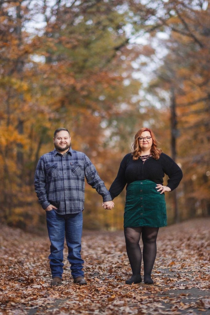 A man and a woman stand holding hands on a leafcovered path in a forest during autumn. The woman has red hair and wears a black top and green skirt, while the man wears a plaid shirt and jeans. The moody dusk light casts an eerie glow over the scene, adding a touch of mystery to their quiet moment.