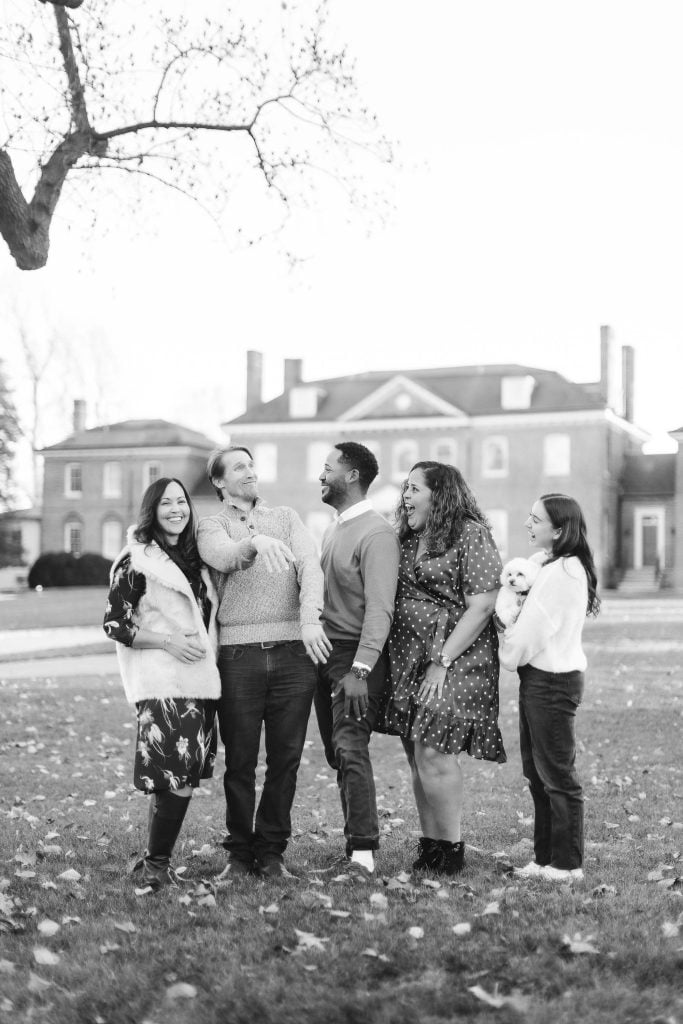 A black and white photo of an extended family, featuring five adults and a child standing together on a lawn, with the Belair Mansion in the background. Some people are laughing, and one person holds a baby.