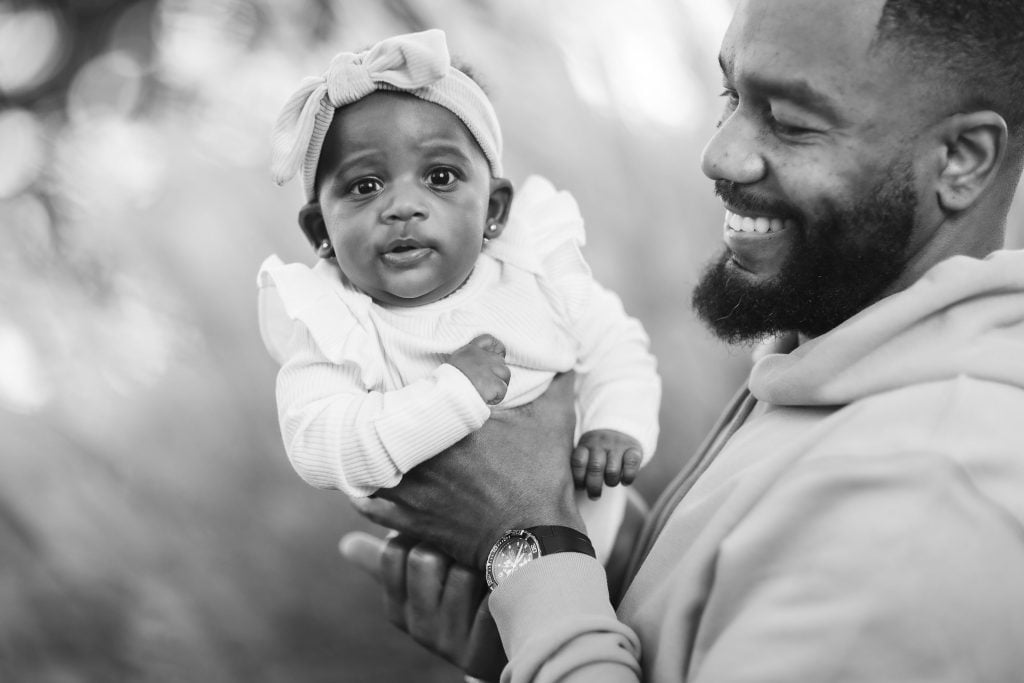 A blackandwhite portrait shows a man joyfully holding a baby, who wears a charming headband with a bow. Both are smiling, capturing a heartfelt family moment.