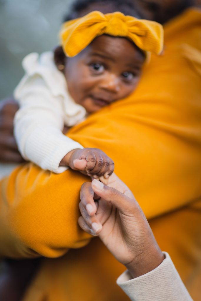 A family portrait captures a baby wearing a yellow bow, held by an adult in a yellow shirt, while another hand gently touches the baby’s hand.