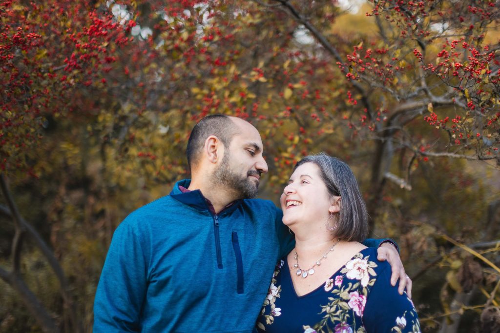 A person in a blue jacket stands with their arm around another person in a floral blouse, both smiling, in front of a tree with red berries at Greenspring Garden in Virginia.