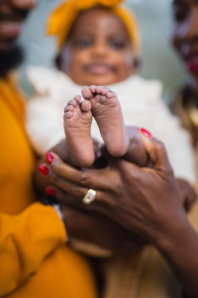 Portrait of a baby's feet held by two adults. The baby, dressed in a white outfit with a yellow headband, is cradled by family members donned in matching yellow attire.