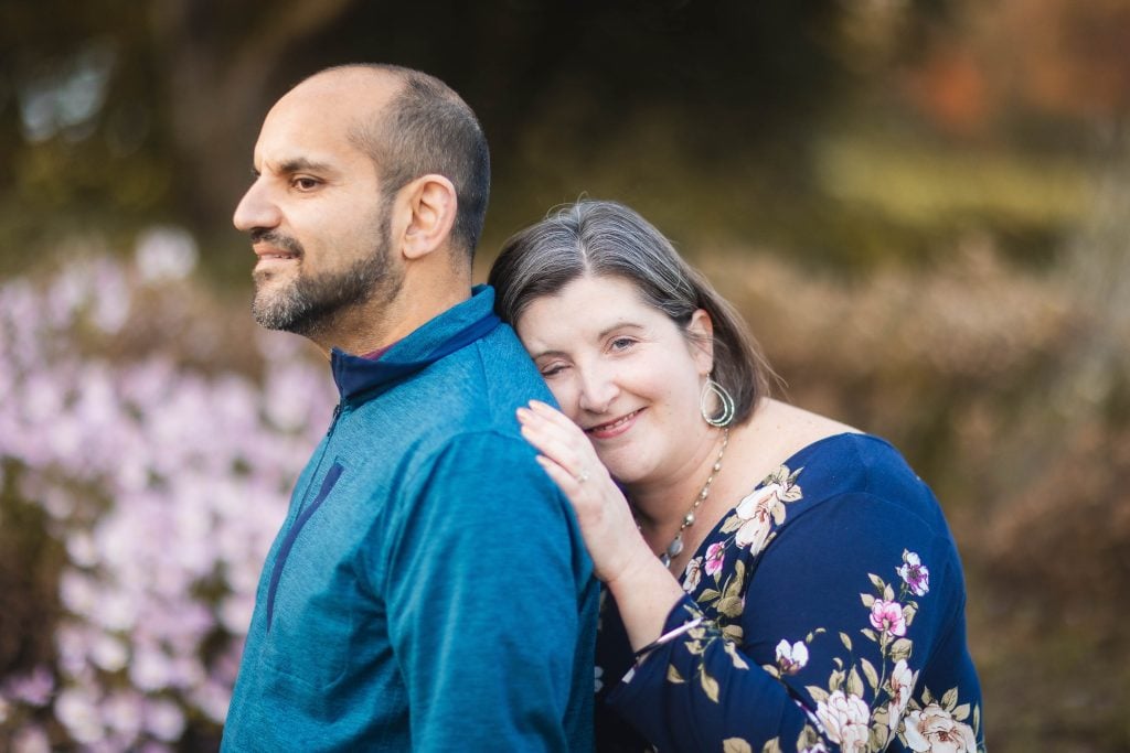 A woman smiles and leans on a man's shoulder from behind. The man looks off to the side. They are standing outdoors in Greenspring Garden, Virginia, with pink flowers in the background.