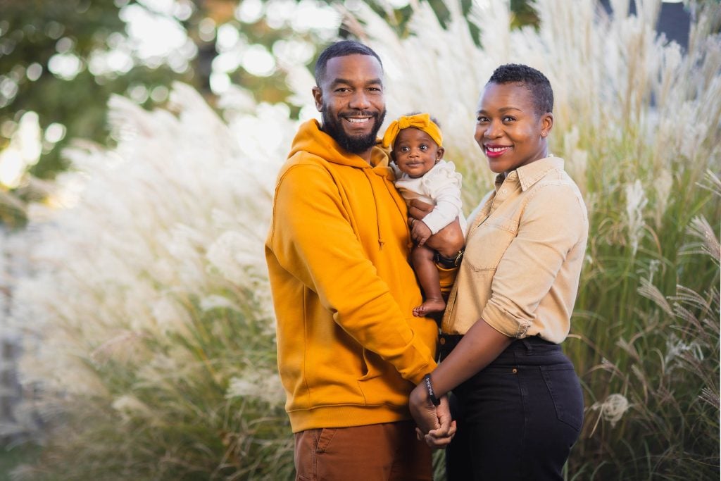 A smiling family stands outdoors, dressed in matching yellow outfits. The portrait captures the couple holding their baby against a backdrop of tall, fluffy grasses.