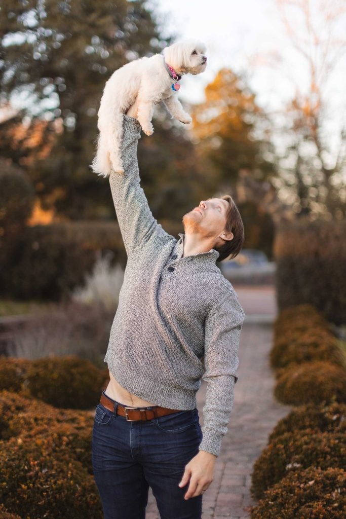 A man joyfully holds a small white dog above his head outdoors on a path lined with bushes, creating a vibrant portrait fit for an extended family album at Belair Mansion.