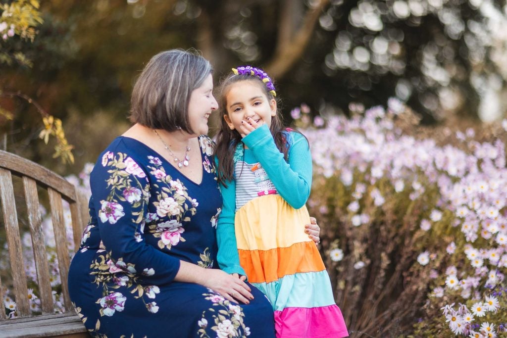 A woman in a floral dress and a young girl in a colorful dress sit on a wooden bench at Greenspring Garden, Alexandria, surrounded by blooming flowers. The girl is covering her mouth while the woman smiles at her.