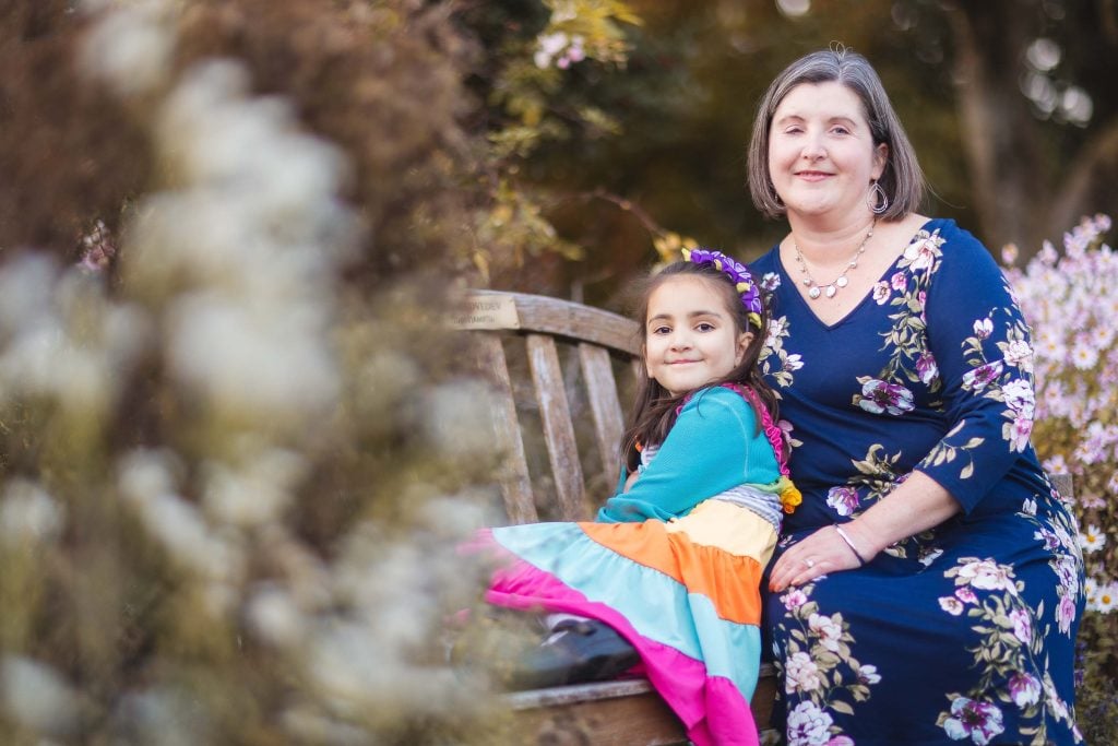 A woman and a child sit together on a wooden bench in Greenspring Garden. The woman wears a dark floral dress, and the child wears a colorful dress. They both smile at the camera, capturing a charming portrait in the heart of Virginia.