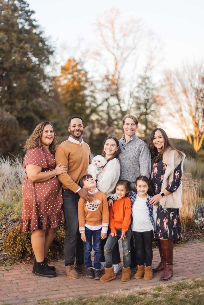 A family of seven people and a small white dog stand together outdoors, smiling at the camera. The background features trees and a clear sky, creating a perfect portrait in front of Belair Mansion.