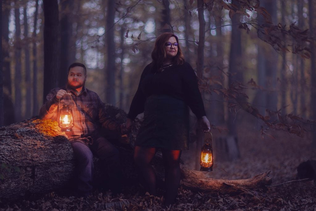 A man and woman hold lanterns while standing and sitting on a fallen tree trunk in a dimly lit forest with autumn leaves, creating a moody portrait at dusk.