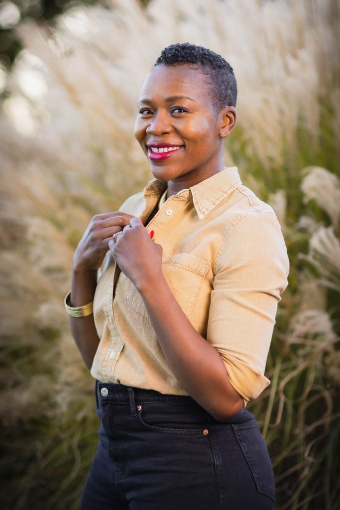 A person with a short haircut smiles while standing outside in front of tall grass, wearing a beige buttonup shirt and dark highwaisted pants, creating a warm portrait that exudes a sense of family connection.