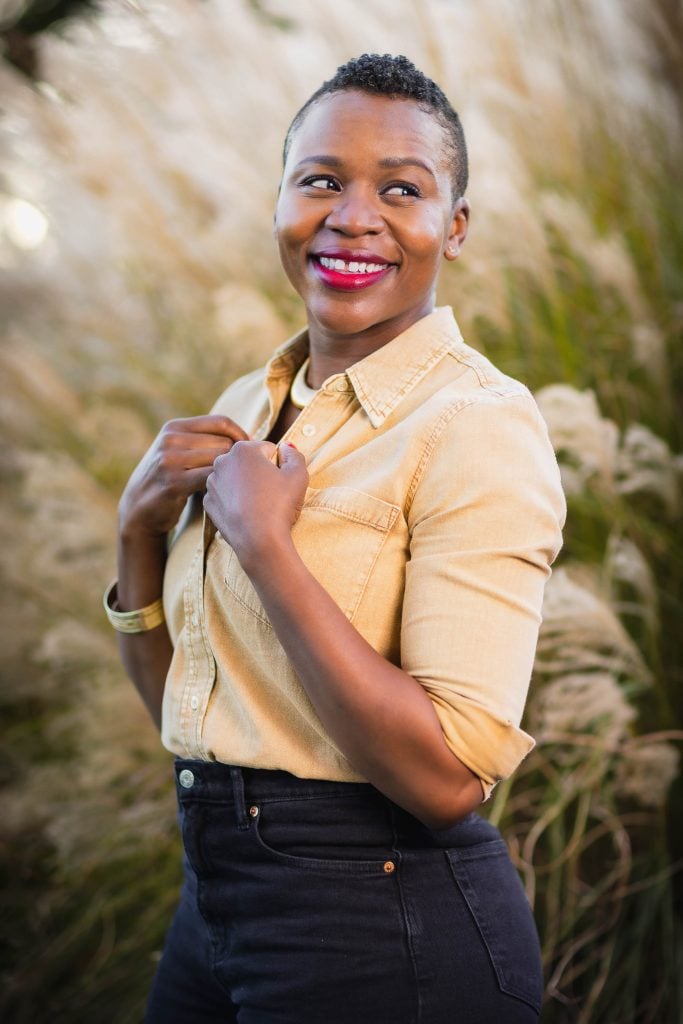 A woman in a yellow buttondown shirt and dark pants smiles while standing in front of tall grass, her hands touching her collar, creating a perfect family portrait.