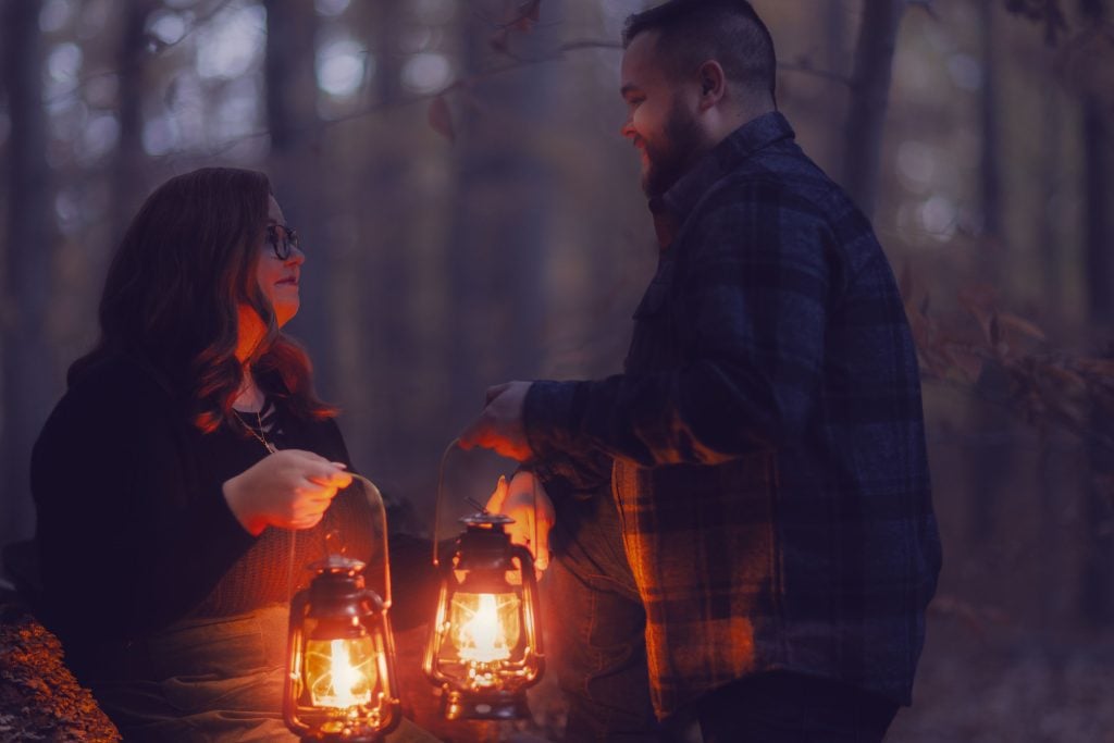 A couple stand in a forest at dusk, each holding a lit lantern. They face each other and smile warmly as the moody light from the lanterns illuminates their faces.