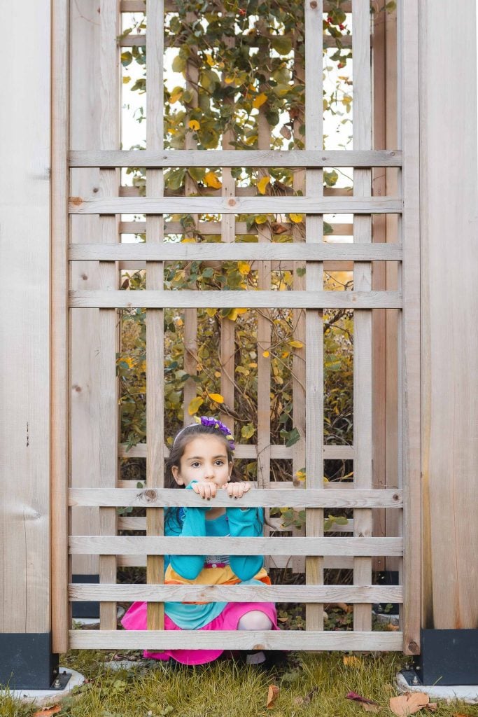 A young girl with a bow in her hair crouches behind a wooden lattice fence, looking through the gaps. She is wearing a blue top and a colorful skirt. Verdant foliage is visible behind her in this charming portrait captured at Greenspring Garden in Virginia.