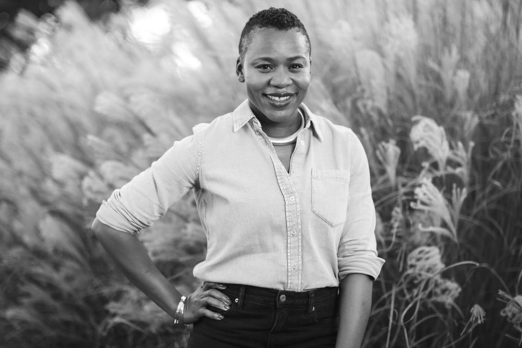 Black and white portrait of a person smiling, standing outdoors with tall grass in the background. They are wearing a lightcolored shirt and dark pants, with one hand on their hip.